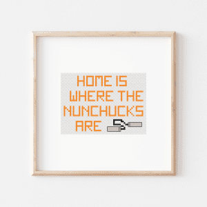 Cross stitch of blocky orange text that reads "Home is where the nunchucks are" with small grey and black nunchucks in the bottom right corner.