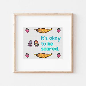Cross stitch featuring sprites from the video game Celeste. There is a strawberry in each corner, a feather on the top and bottom between strawberries, and in the middle, a sprite of Badeline facing Madeline. There is light blue left-justified text reading "It's okay to be scared."